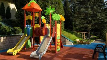 A playground with slides and climbing structures, surrounded by trees and a building in the background.