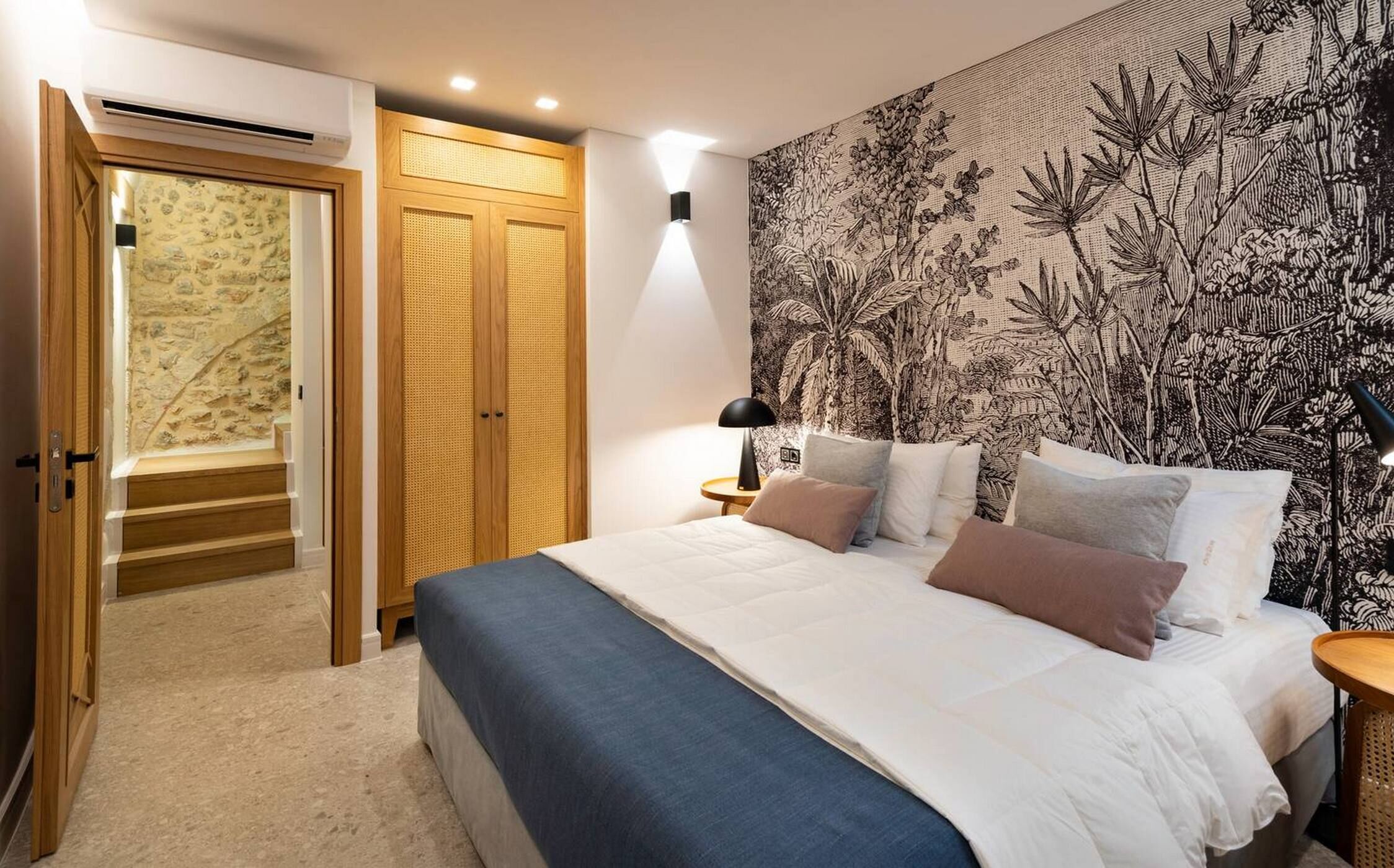 A bedroom with a bed, a nightstand, a lamp, a wardrobe, and a wall with a botanical pattern.