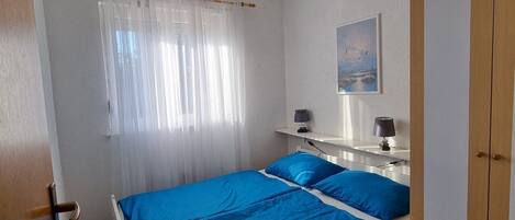 A bedroom with a bed, a bedside table with a lamp, a wardrobe, and a window with sheer curtains.