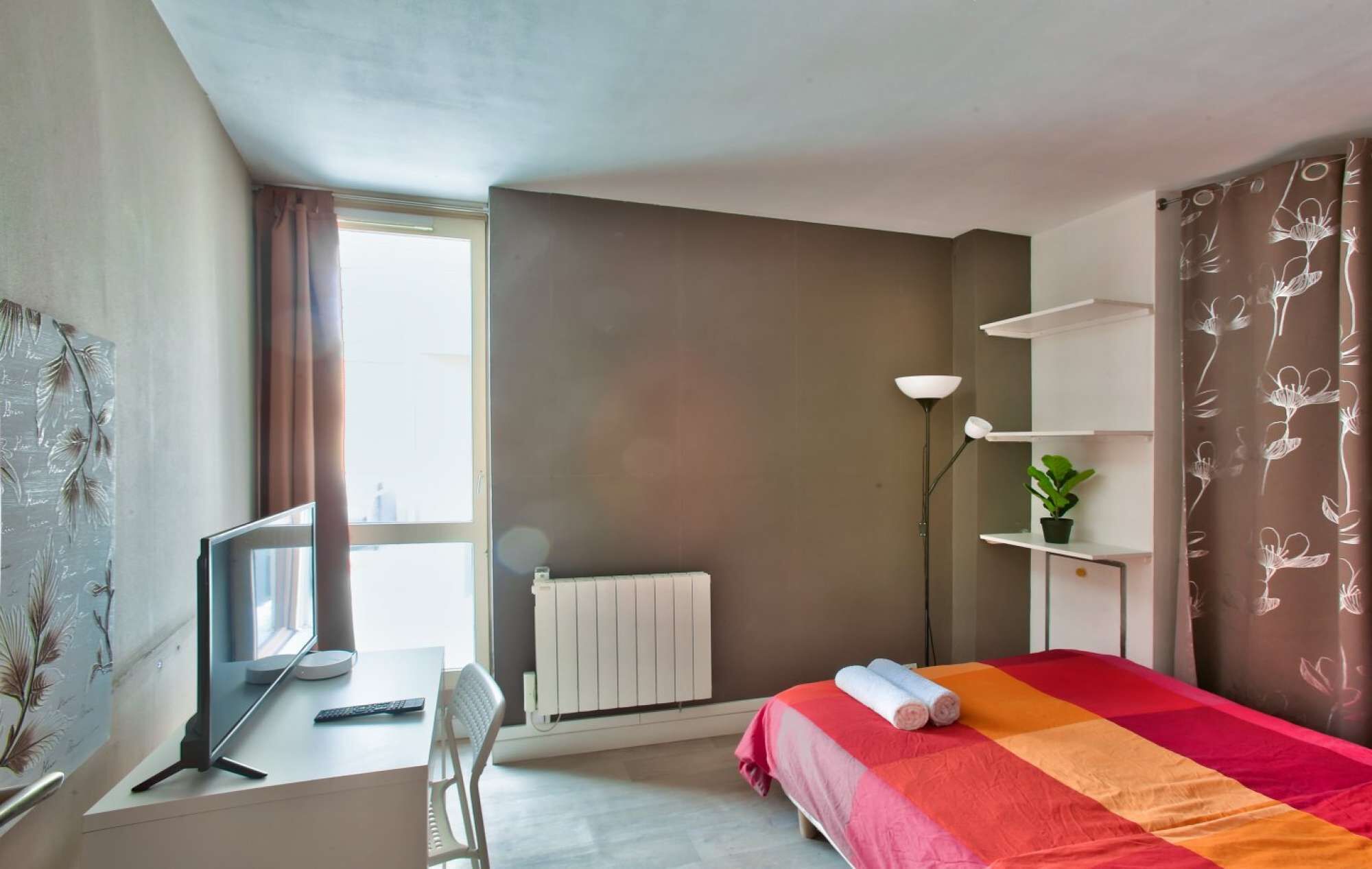 A modern hotel room with a desk, chair, bed, radiator, and a plant.