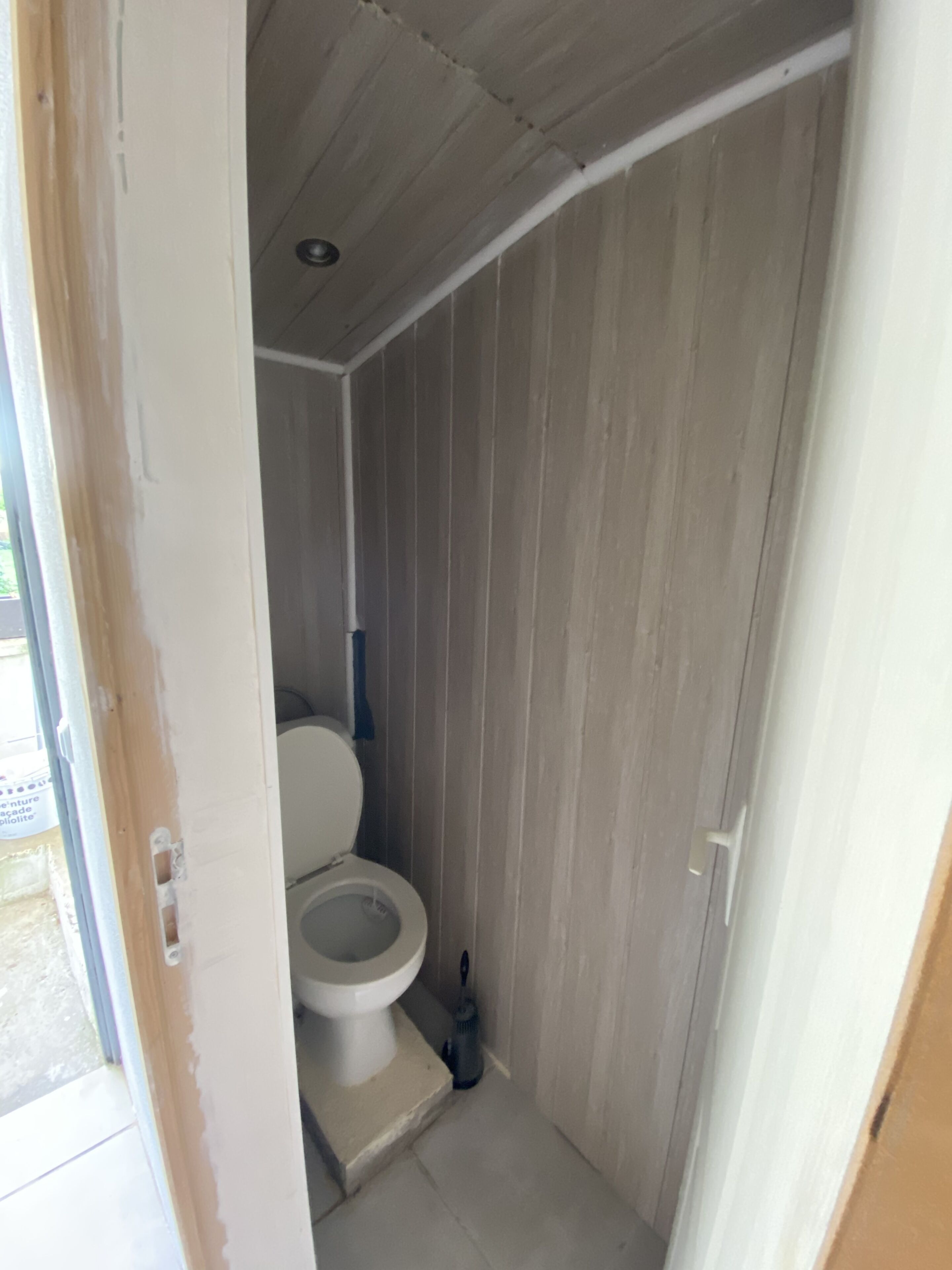 Une petite salle de bain étroite, équipée de toilettes et d’un sèche-mains. Les murs sont recouverts de lambris en bois, et une porte donne accès à une autre pièce.