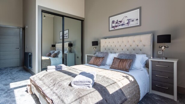 A bedroom with a large bed, a wardrobe, a bedside table with a lamp, and a framed picture on the wall. - Trafalgar Lane (Edinburgh)