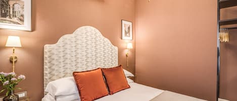 A hotel room with a large bed, two pillows, a bedside table with a lamp, a vase with flowers, and a chandelier.