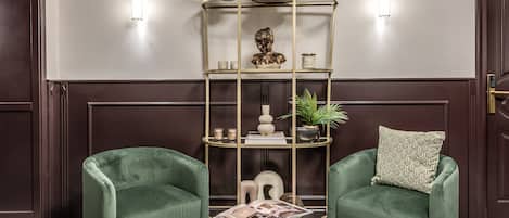 A room with two green armchairs, a round gold-framed side table, and a gold-framed shelving unit with decorative items.