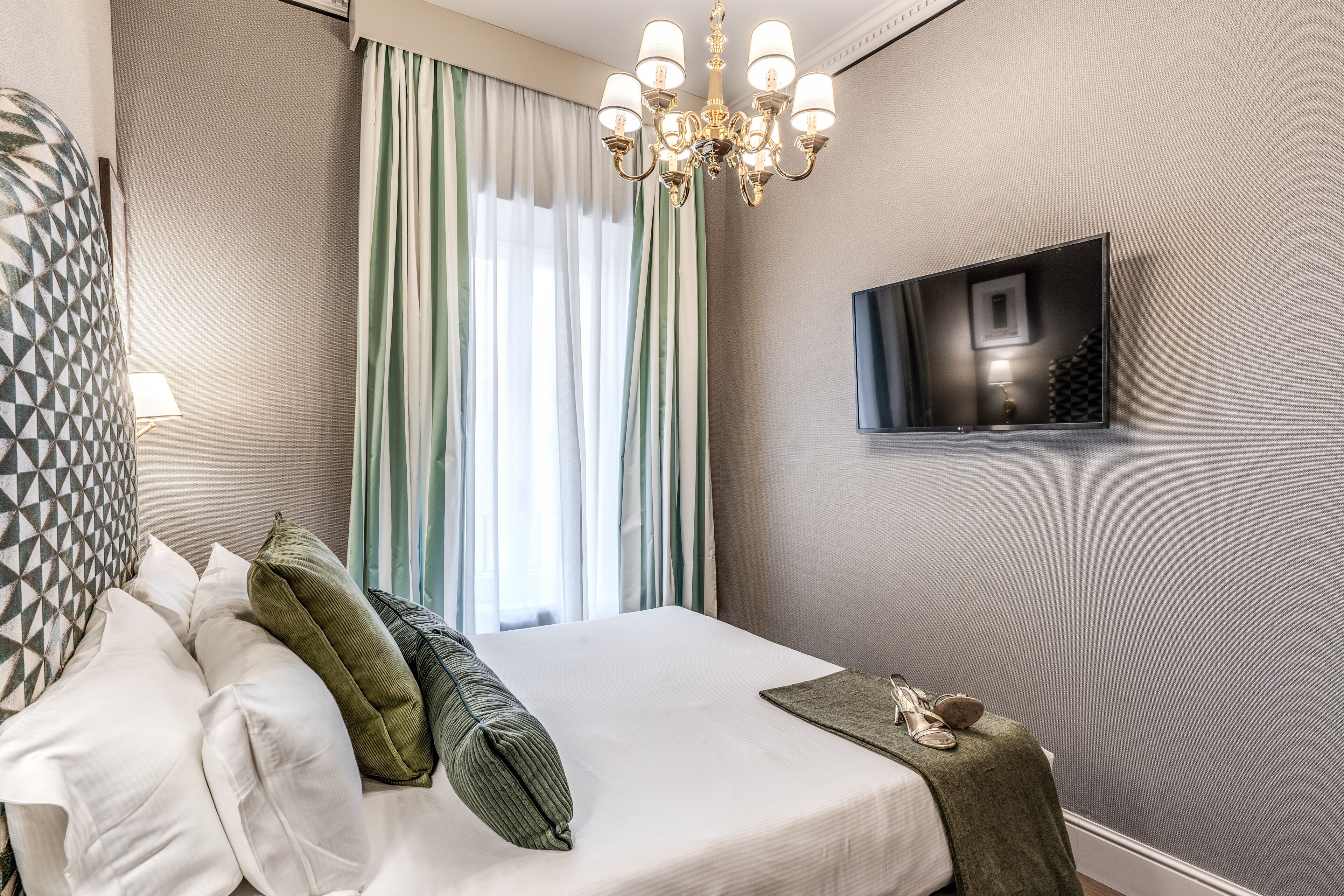 A hotel room with a bed, pillows, a TV mounted on the wall, and a window with curtains.