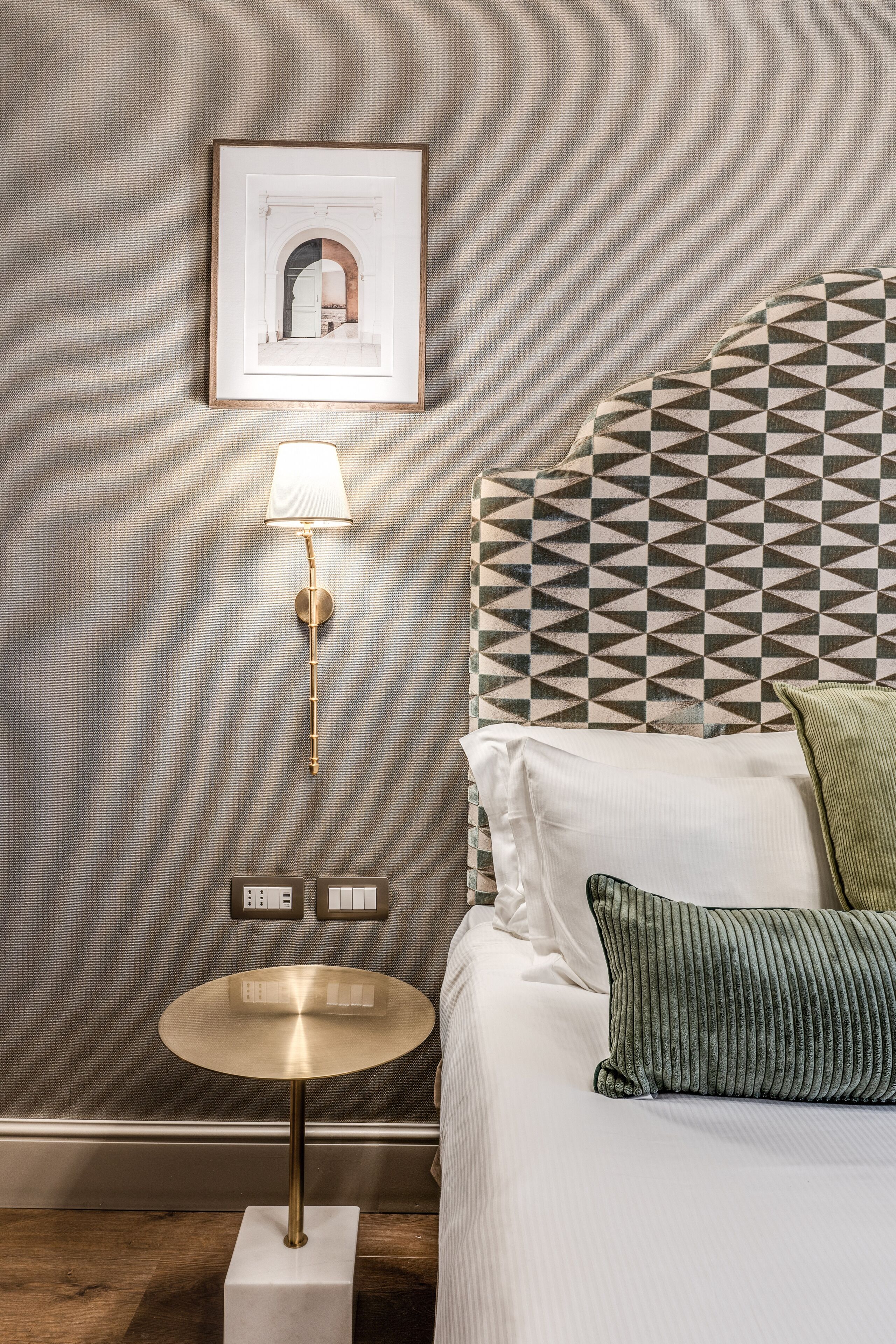 Une chambre d’hôtel bien rangée, avec une tête de lit à motifs, une lampe de chevet, une table ronde et un tableau encadré au mur.