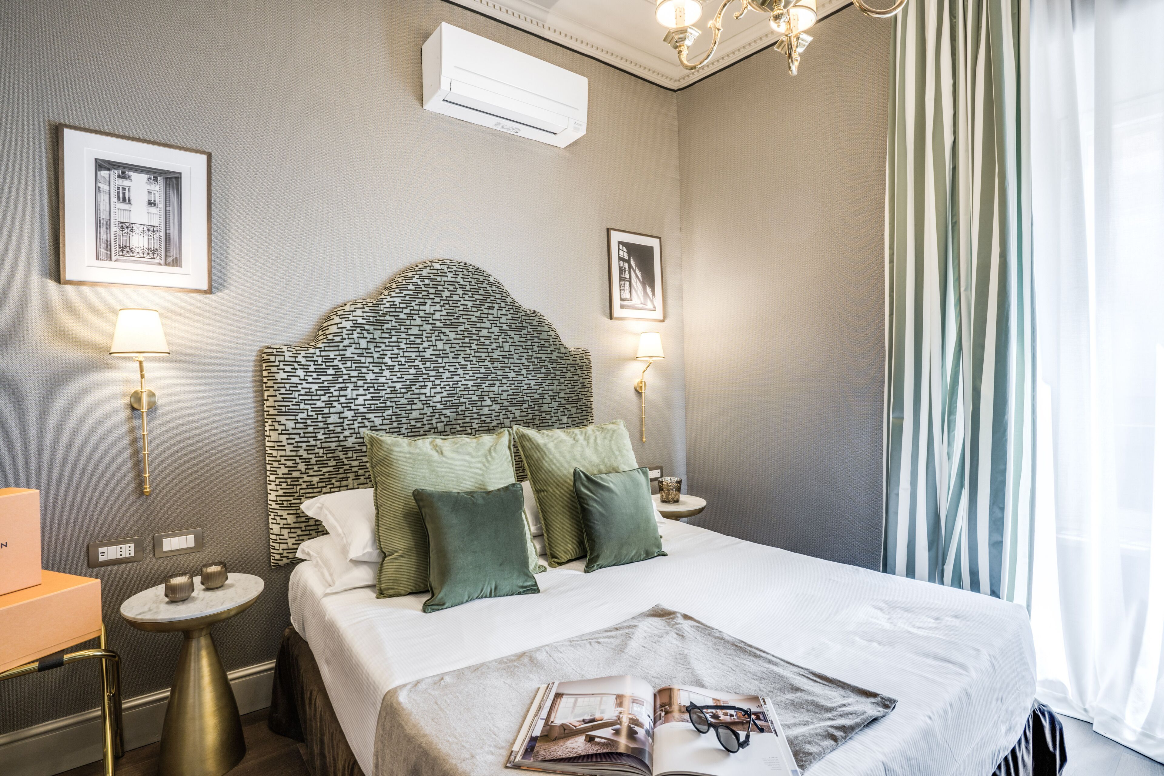 Habitación de hotel ordenada con un cabecero estampado, almohadas verdes, una mesita de noche con lámpara y una revista sobre la cama.