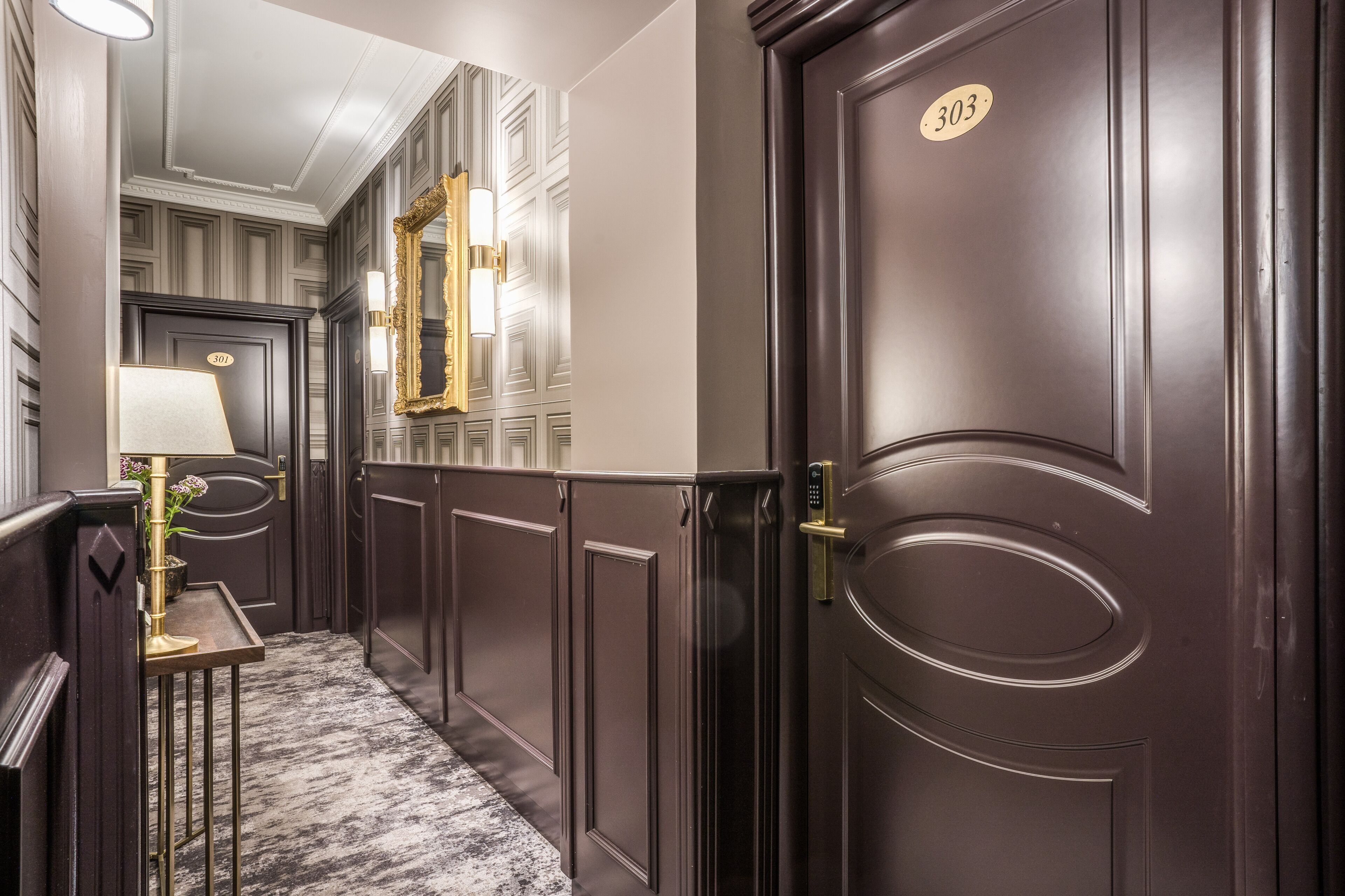 A hallway with a dark wooden door marked with the number 303, a wall-mounted mirror, and a small table with a lamp.