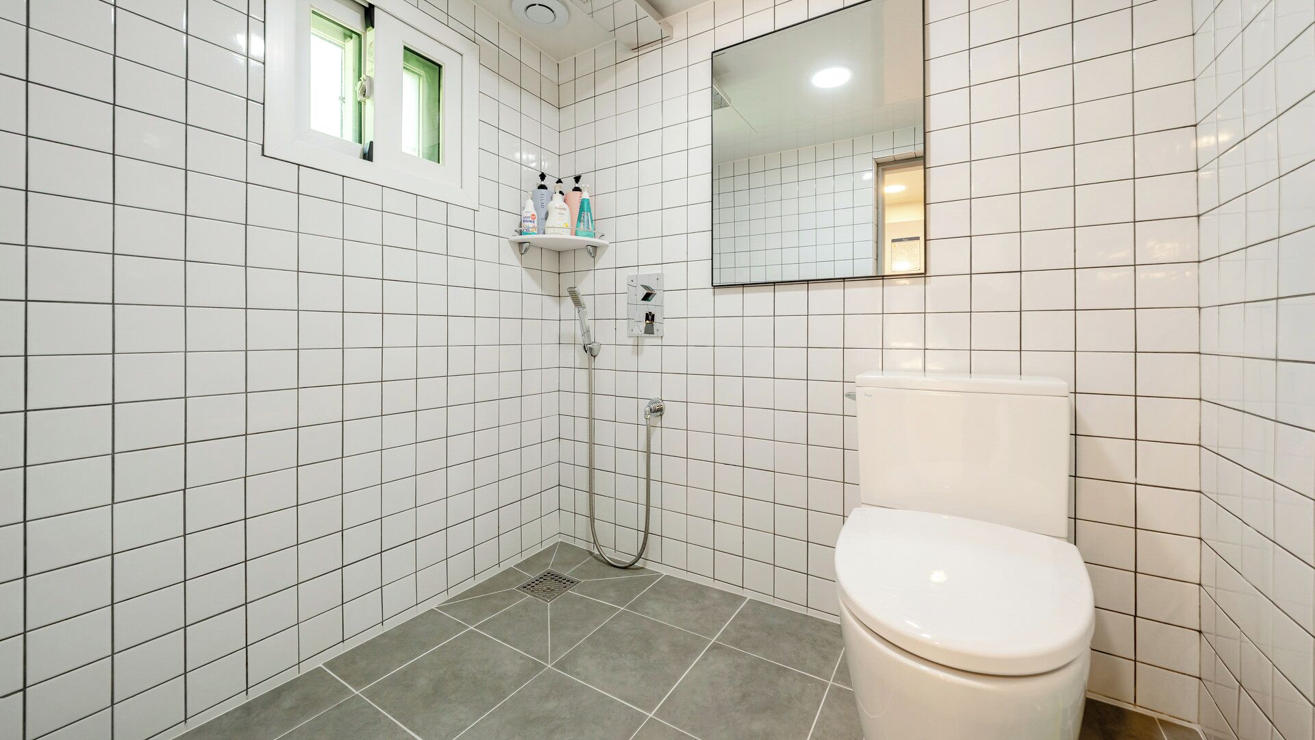 A bathroom with white tiled walls, a toilet, a shower with a handheld showerhead, and a window with a view.
