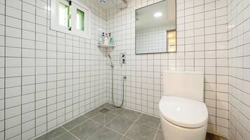 A bathroom with white tiled walls, a toilet, a shower with a handheld showerhead, and a window with a view.