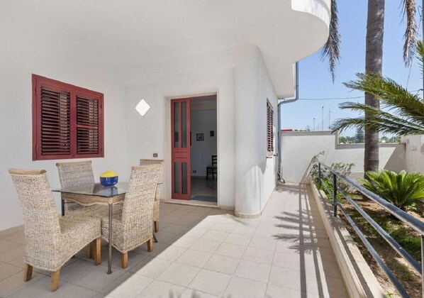 A balcony with wicker furniture, a glass table, and a red door leading to an interior space.