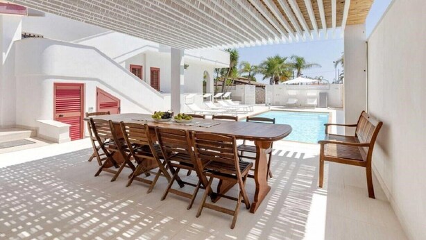Un espace aménagé au bord de la piscine, avec une table et des chaises en bois, une piscine et un coin salon abrité.