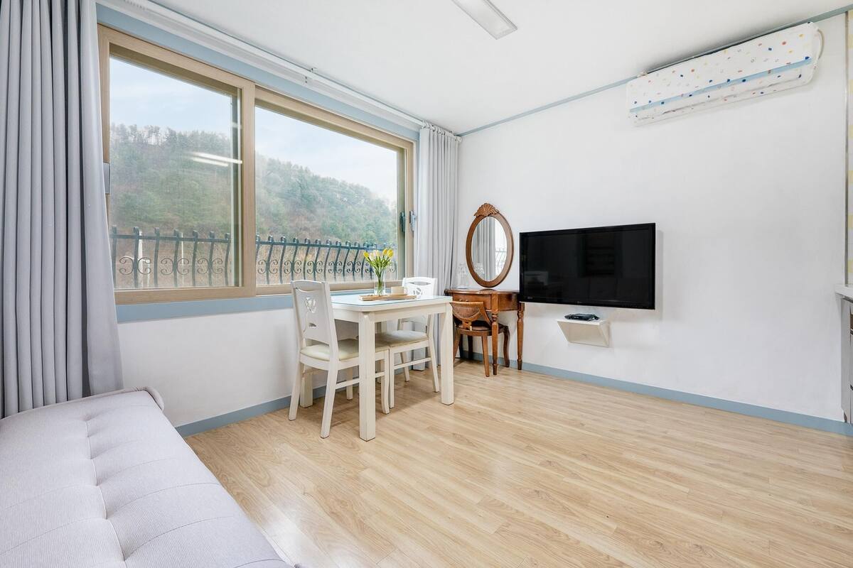A modern hotel room with a sofa, a dining table with chairs, a TV, and a window with a view of trees.
