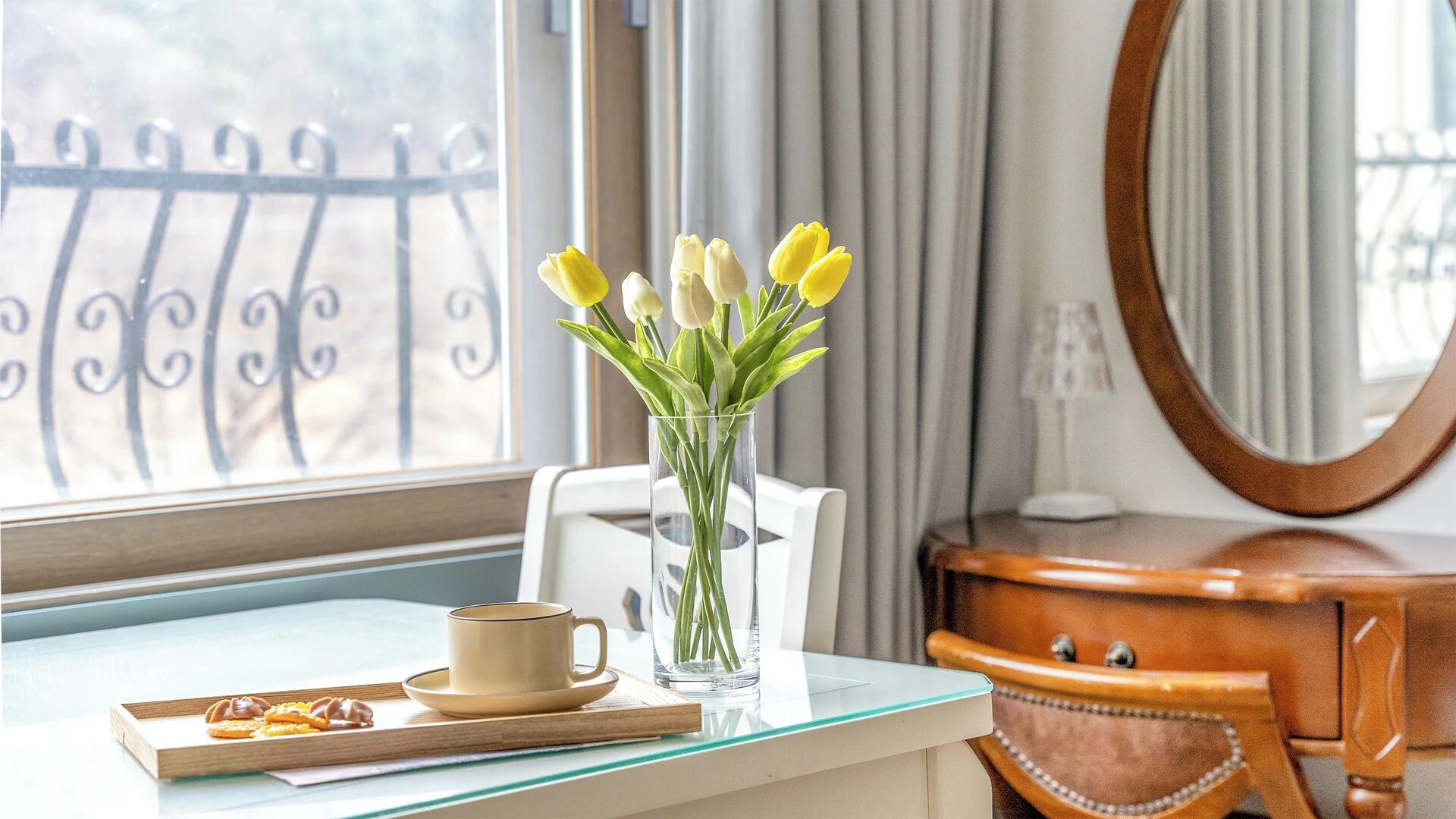 A hotel room with a dining table, a vase with yellow flowers, a cup, and a tray with pastries.
