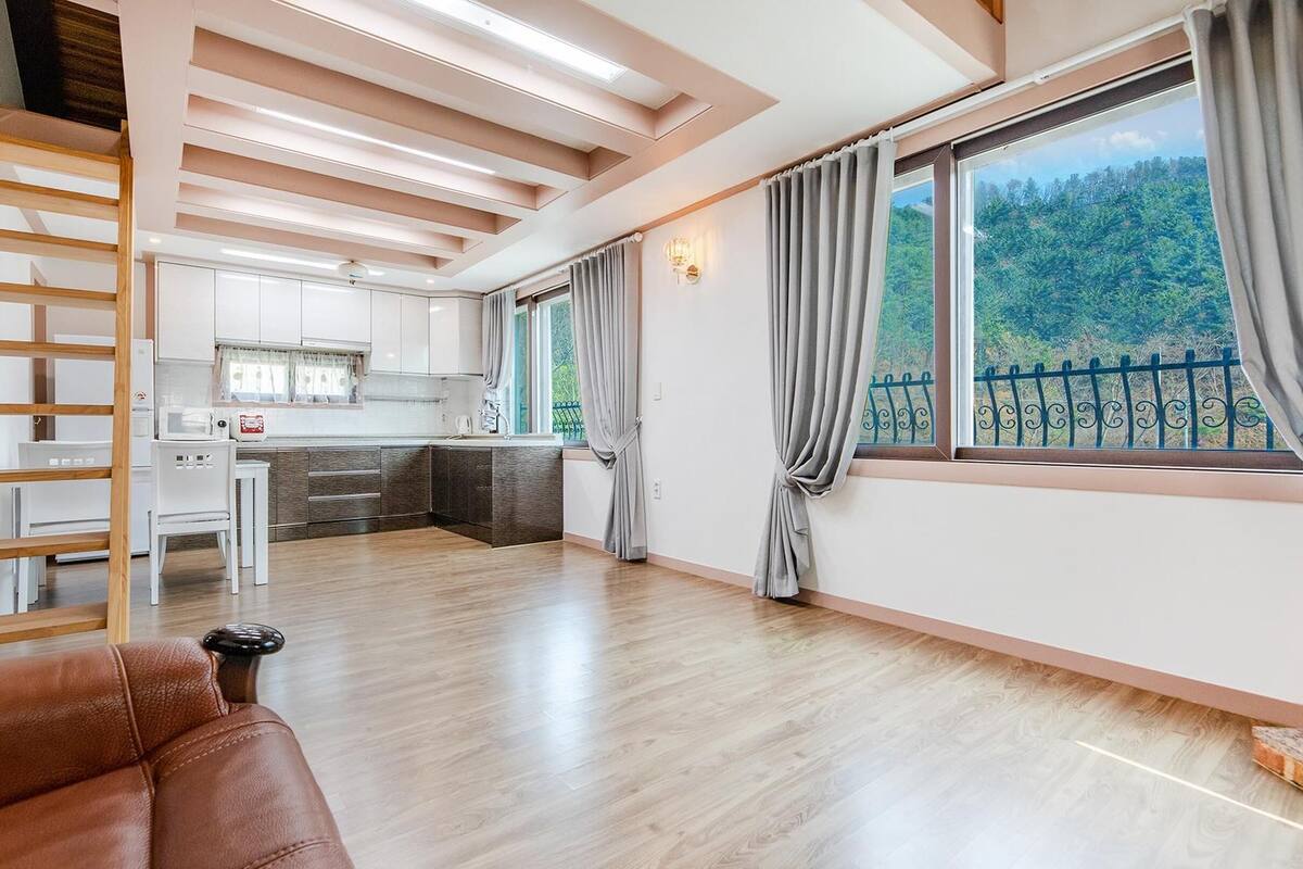 A spacious living area with a wooden floor, a brown leather sofa, a kitchen with white cabinets, and a balcony with a view of greenery.