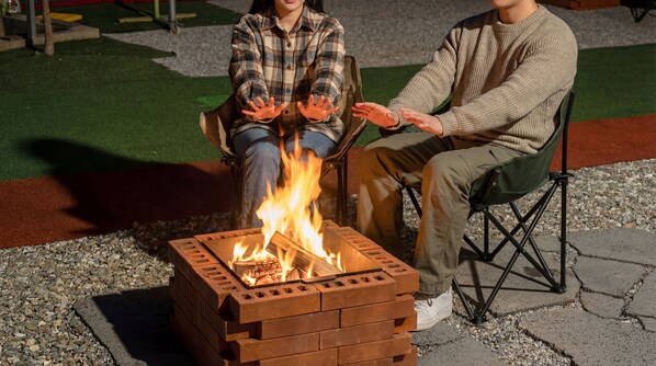 Two people sitting by a fire pit with a brick design, outdoors on a paved area with gravel and grass.