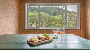 A table with a cheese board, grapes, and wine glasses, with a window view of a forested landscape.