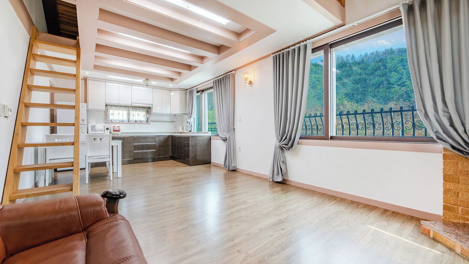 A spacious living area with a wooden floor, a brown leather sofa, a kitchen in the background, and a large window with curtains.