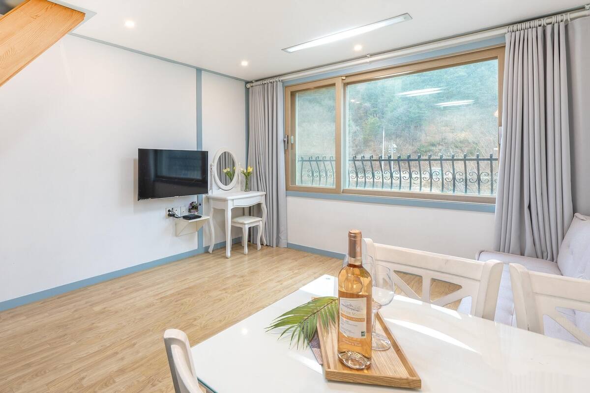 A modern room with a wooden ceiling, a flat-screen TV mounted on the wall, a small white vanity with a mirror, a dining table with a bottle of wine and a tray, and a large window with curtains.