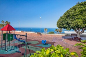 Une aire de jeux avec des balançoires et un toboggan, des bancs, un arbre et une vue sur la mer.