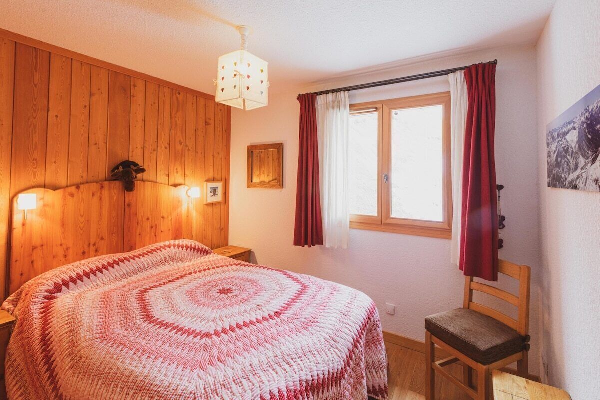 A bedroom with a wooden headboard, a bed with a patterned blanket, a wooden chair, a window with red curtains, and a wall-mounted light fixture.