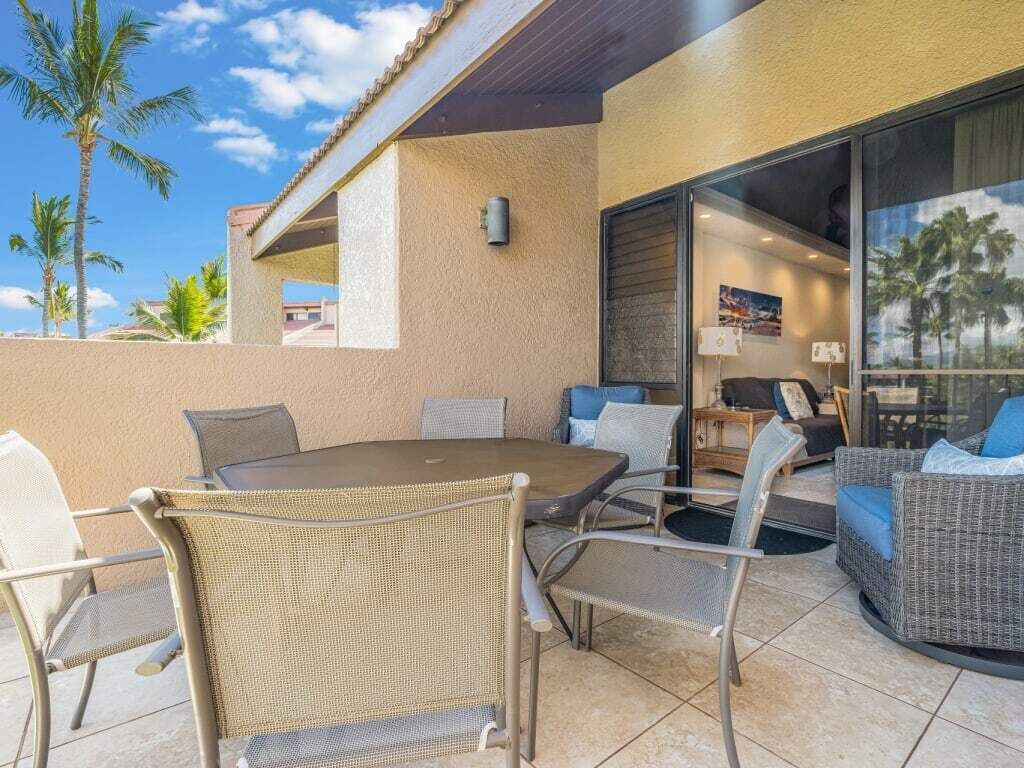 An outdoor patio with a round table and wicker chairs, a glass door leading inside, and a view of palm trees.