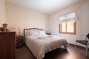 Une chambre à coucher comprenant un lit, une commode, une chaise, une fenêtre avec des stores et une table de chevet avec une lampe.