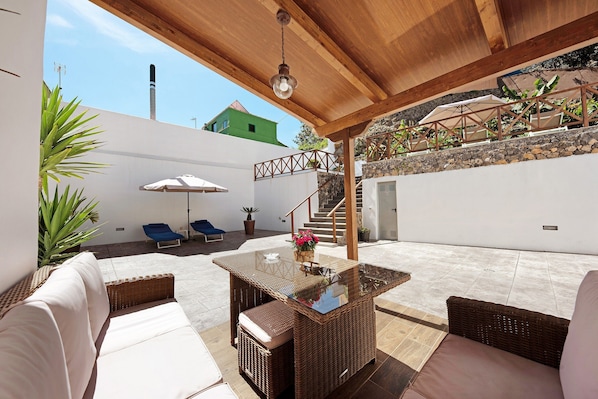 Une terrasse extérieure avec des meubles en osier, une table à plateau en verre et un luminaire suspendu au plafond.
