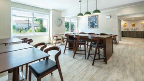 A spacious dining area with wooden tables and chairs, large windows offering a view of the outside, and pendant lights hanging from the ceiling.