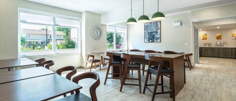 A spacious dining area with wooden tables and chairs, large windows offering a view of the outside, and pendant lights hanging from the ceiling.