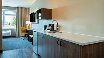 Une cuisine d’hôtel moderne avec un plan de travail blanc, des meubles en bois foncé, un téléviseur encastré et une fenêtre avec des rideaux.