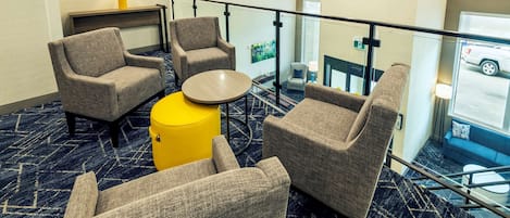 A hotel lobby with four grey armchairs, a yellow ottoman, and a round table. A glass railing overlooks a parking area.