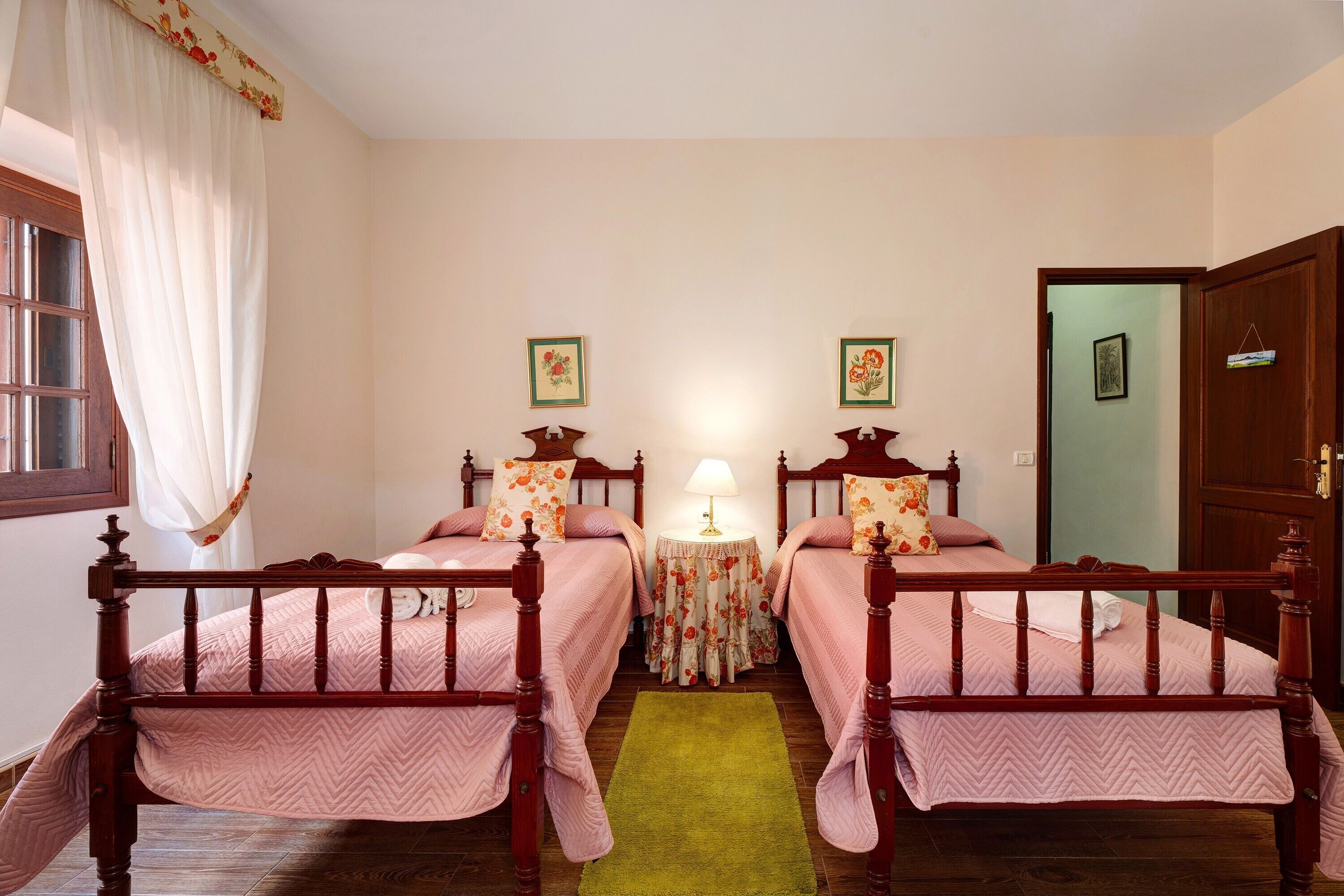 Una habitación con dos camas de madera, un tapete verde, una mesita redonda con una lámpara y una puerta.