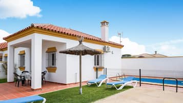 A white building with a red-tiled roof, a swimming pool, and blue lounge chairs.