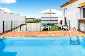 Une piscine avec une terrasse en bois, un parasol en chaume et deux chaises longues.
