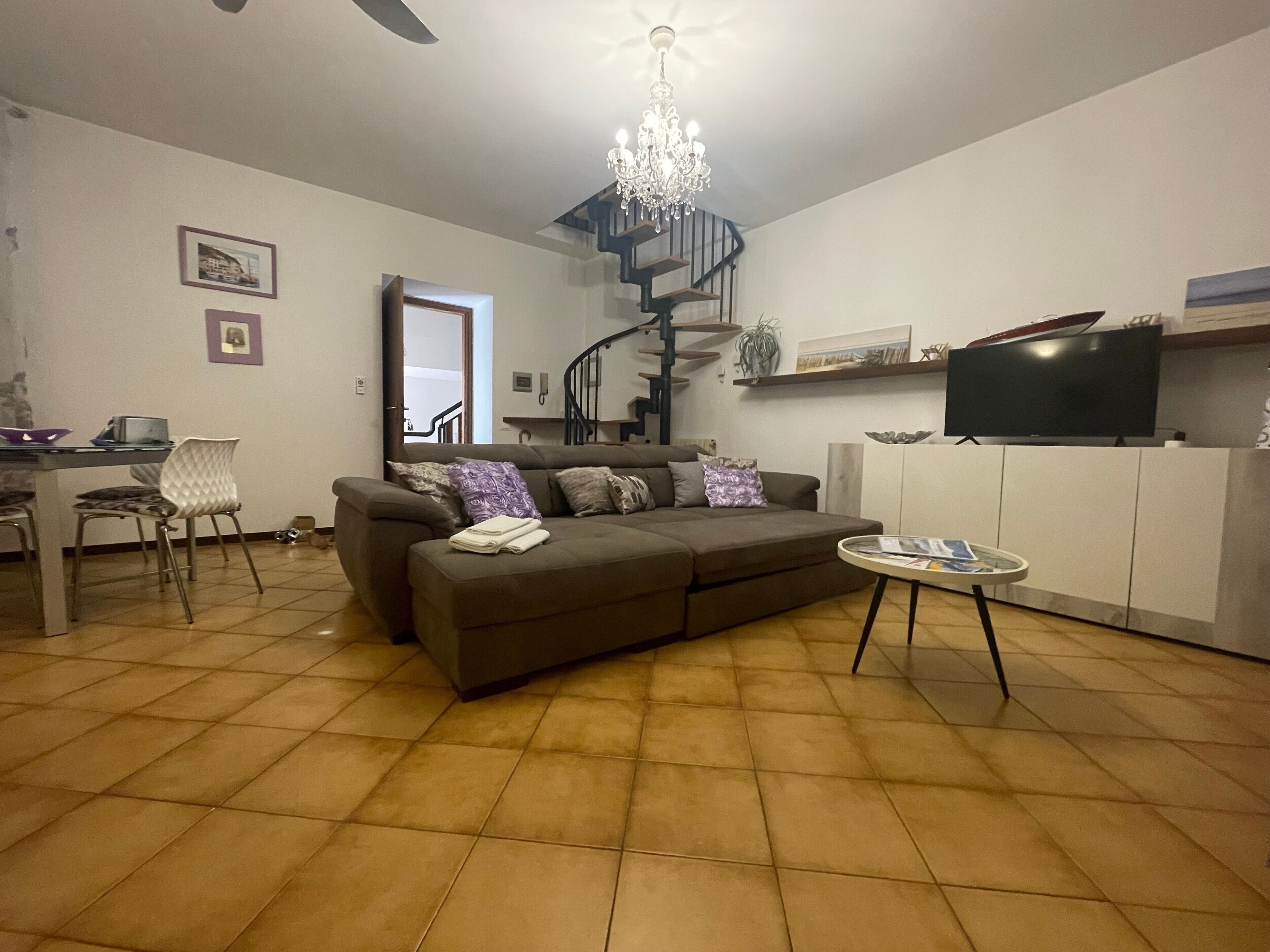 Ein Wohnzimmer mit einer Couch, einem Esstisch, Stühlen, einer Chandelier, einer Treppe und einem Fernseher.