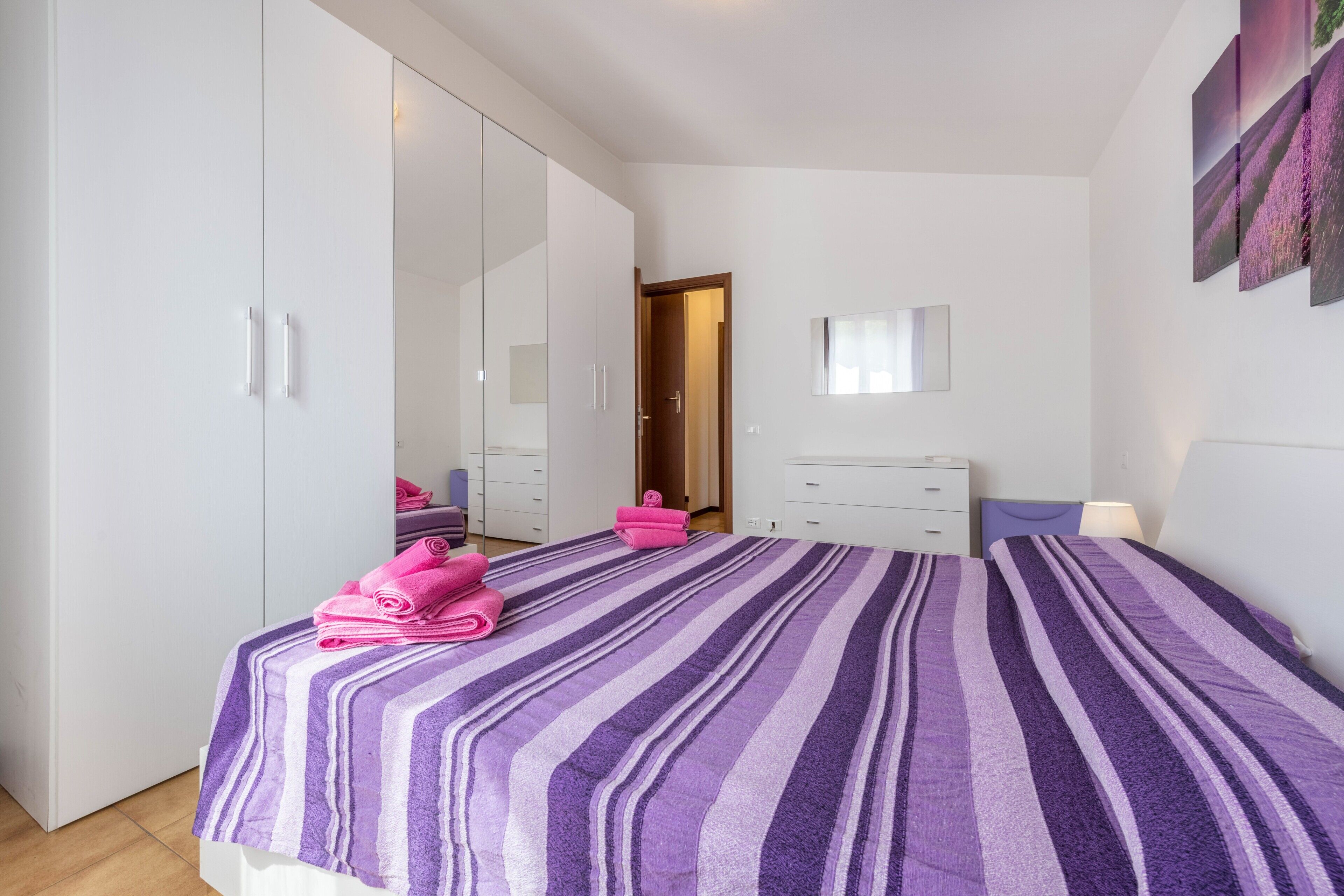 A bedroom with a large mirror, a bed with striped bedding, a wardrobe, and a bedside table with a lamp.
