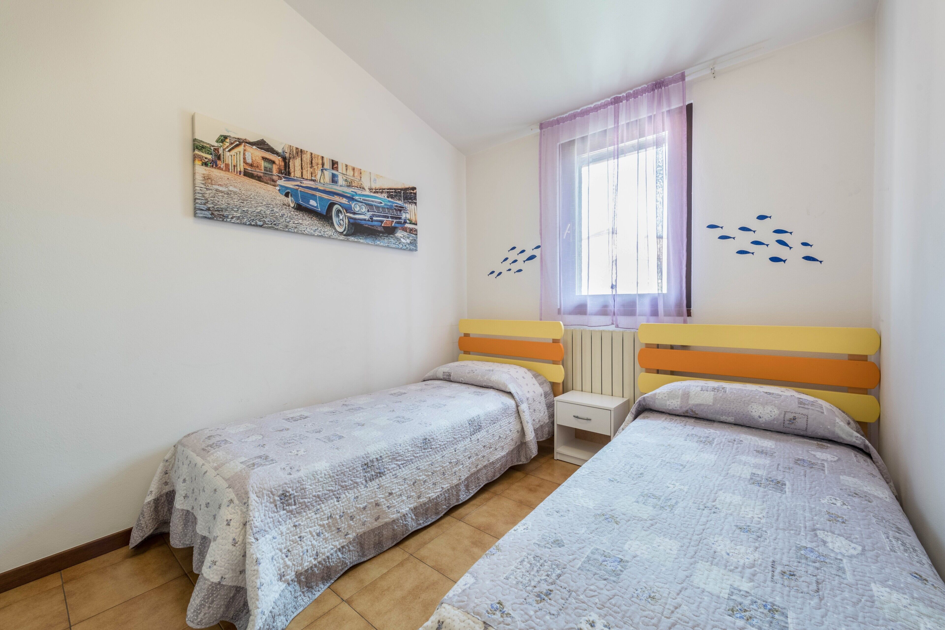 A bedroom with two beds, a window with curtains, a wall-mounted painting, and a small bedside table.