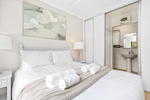 Une chambre d’hôtel comprenant un lit, une table de chevet, une lampe, un tableau et une salle de bain avec un lavabo et un miroir.