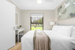 A hotel room with a bed, a bedside table, a lamp, a window with a view of greenery, and a ceiling fan. - Laguna Beach B8 (Cape Town)