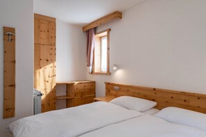 A bedroom with a wooden headboard, a wooden wardrobe, a small window with a checkered curtain, and a radiator. - Rodella A Parte Campitello (Campitello di Fassa)