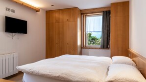 A bedroom with a bed, wooden wardrobe, a TV mounted on the wall, a window with a view, and a radiator.