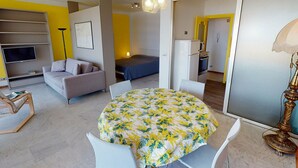 A modern living room with a sofa, a dining table with a floral tablecloth, a TV, and a kitchen area with a refrigerator and microwave. - Spacious Studio with Sea View, Terrace, Near Center and Beaches in Menton (Menton)