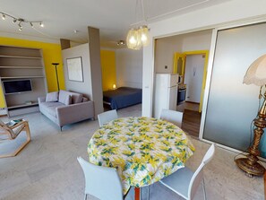A modern living room with a sofa, a dining table with a floral tablecloth, a TV, and a kitchen area with a refrigerator and microwave.