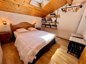 A bedroom with a wooden ceiling, a bed with white bedding, a nightstand, a shelf with storage boxes, and a hanging rack with clothes. - Family apartment with pools in Samoëns (Samoëns)