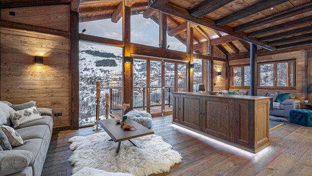 Una cabaña amplia con un interior de madera, un espacio de estar acogedor con un sofá y una mesa de comedor, y vistas a montañas nevadas a través de grandes ventanales.