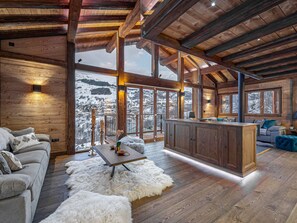 Una cabaña amplia con un interior de madera, un espacio de estar acogedor con un sofá y una mesa de comedor, y vistas a montañas nevadas a través de grandes ventanales.