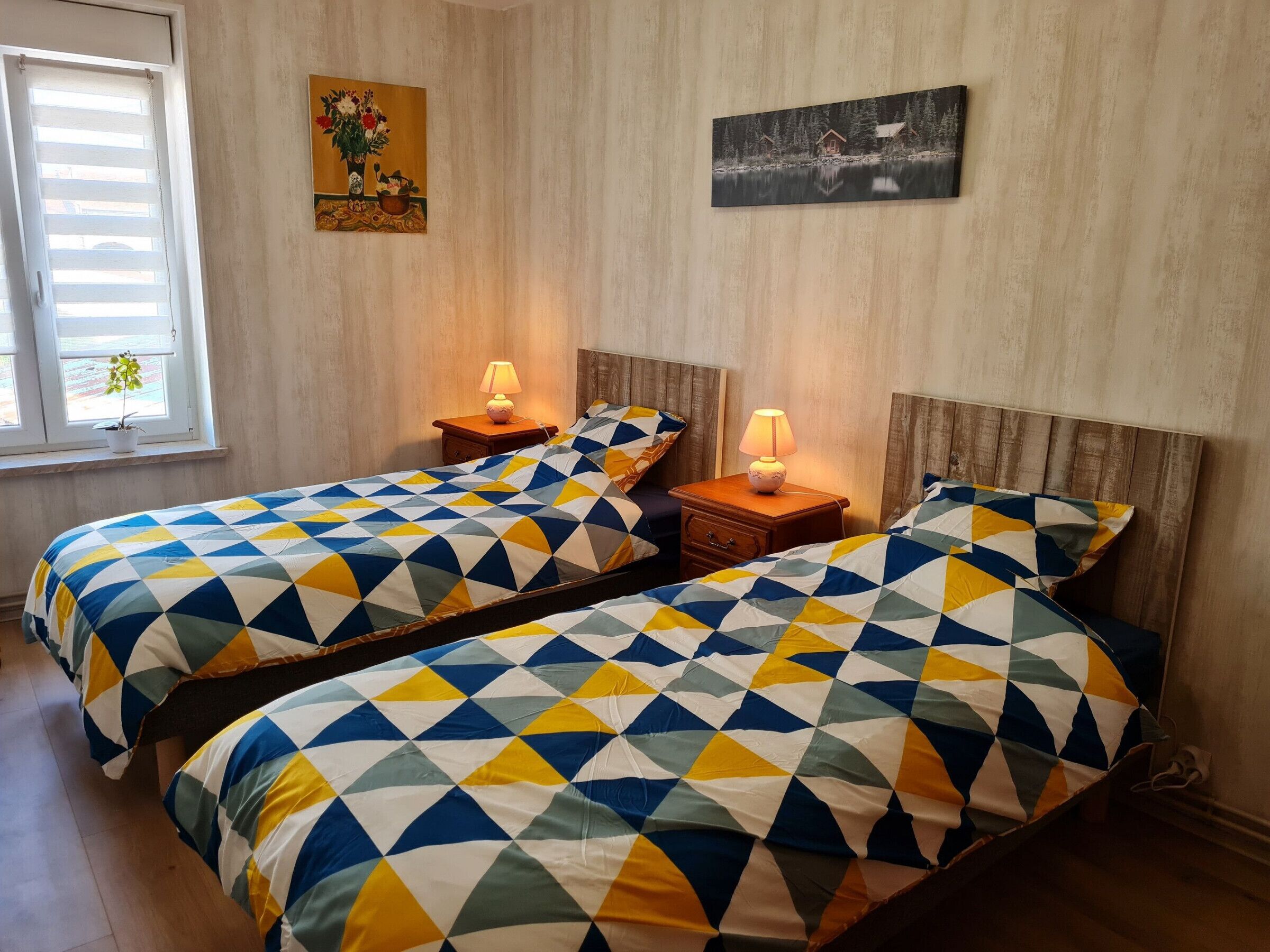 Ein Zimmer mit zwei Betten, jeweils mit einem Bettbezug mit geometrischem Muster, einem Nachttisch mit Lampe, einem Fenster mit Jalousien und zwei an der Wand hängenden Bildern.