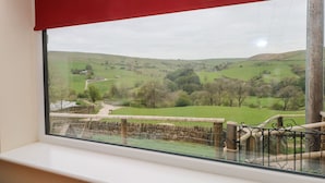 Une fenêtre avec un store rouge, donnant sur un paysage rural avec des champs verdoyants, des murs de pierre et une clôture en bois.