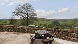 Une table de pique-nique avec des bancs, située dans un cadre rural, entourée de murs en pierre et offrant une vue sur des collines ondoyantes et un arbre.